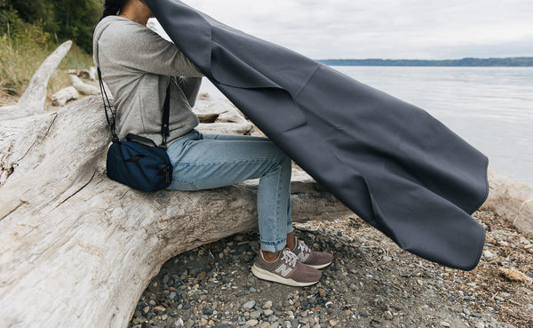 A person sitting on a log at the beach getting ready to lay a Large Shepherd's Wool Utility Cloth in Matter Grey on their lap.